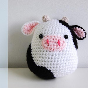 May include: A handmade crochet cow with a white body, black patches, and pink accents on the ears and snout. The cow has black eyes and small beige horns. The word "PATTERN" is written vertically in black letters on the left side.