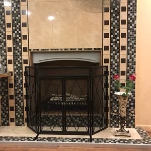 Puede incluir: Una pantalla de chimenea de metal negro con un patrón de diamante, delante de una chimenea con una repisa blanca y una pared de azulejos beige. La pared tiene un reloj decorativo con una esfera plateada y agujas negras. Hay rosas rojas en un jarrón en el lado derecho de la chimenea.