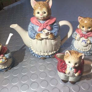 Op de afbeelding: Een set van vier keramische kattenfiguren, waaronder een theepot, een roomkan, een suikerpot en een klein kopje. De figuren zijn allemaal gekleed in blauw-witte kleding met roze accenten. De theepot heeft een tuit en een handvat, en de roomkan en de suikerpot hebben handvatten.