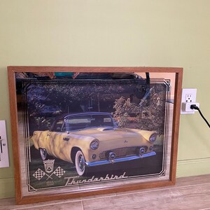 May include: A framed mirror with a vintage image of a yellow 1955 Ford Thunderbird convertible. The mirror has a wooden frame and the image is surrounded by a silver border. The text "Thunderbird" and "1955" are printed below the car.