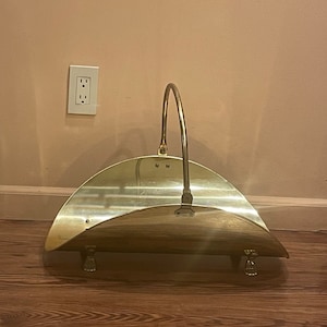 May include: A gold metal log holder with a curved design and a handle. The holder has two decorative feet and is sitting on a wooden floor.