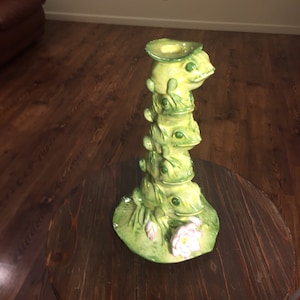 May include: A stack of four green ceramic frogs with a flower on the bottom frog. The frogs are stacked on top of each other and the top frog has a hat on its head. The frogs are on a wooden table.