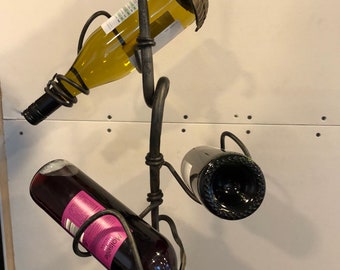 Pode incluir: Um suporte de vinho de metal preto com três garrafas de vinho. O suporte está pendurado no teto e tem um design de folha. As garrafas são um vinho branco, um vinho tinto e um vinho rosé.