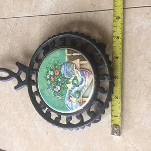 Puede incluir: Posavasos de hierro fundido negro con una baldosa redonda de cerámica verde y blanca con un bodegón de frutas y flores.