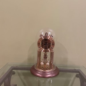 May include: A gold-toned clock with Roman numeral markings on the face, encased in a glass dome. The clock has a pendulum and weights, and sits on a glass table.