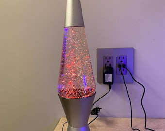 May include: A silver glitter lava lamp with red and blue lights. The lamp is plugged into an electrical outlet.
