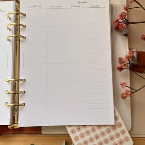 Puede incluir: Una agenda con un dise&ntilde;o de calendario semanal, con encabezados para viernes, s&aacute;bado, domingo y notas. Se incluye una hoja de pegatinas redondas con fechas. La agenda est&aacute; abierta, revelando los anillos dorados y una combinaci&oacute;n de colores neutros.