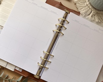 May include: Open planner with a monthly layout. The pages are white with a grid pattern and headings for days of the week. The planner has a gold-colored ring binder and a brown leather cover. A pen rests on the wooden surface.