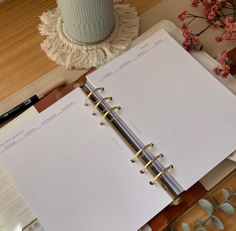 Puede incluir: Planificador abierto con p&aacute;ginas de calendario mensual. El planificador est&aacute; abierto a una vista mensual con l&iacute;neas de cuadr&iacute;cula para cada d&iacute;a. El planificador tiene anillos dorados y una cubierta de cuero marr&oacute;n claro. Un bol&iacute;grafo es visible.