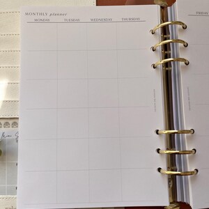 Puede incluir: Agenda abierto en una p&aacute;gina mensual. La p&aacute;gina es blanca con una cuadr&iacute;cula para entradas diarias y encabezados para lunes a jueves. El planificador est&aacute; en una carpeta de cuero con anillos dorados.