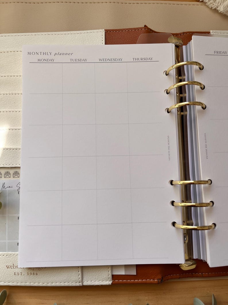 Puede incluir: Agenda abierto en una p&aacute;gina mensual. La p&aacute;gina es blanca con una cuadr&iacute;cula para entradas diarias y encabezados para lunes a jueves. El planificador est&aacute; en una carpeta de cuero con anillos dorados.