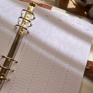 Op de afbeelding: Open planner met goudkleurige metalen ringen en gelijnde pagina's voor maandelijkse en jaarlijkse planning. De pagina's zijn wit met data en maanden in zwart gedrukt. De planner is open voor de maanden maart, april, mei, juni, juli en augustus.