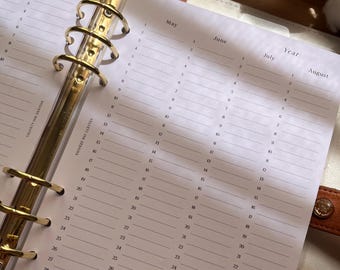 Puede incluir: Agenda abierta con anillas metálicas doradas y páginas rayadas para la planificación mensual y anual. Las páginas son blancas con fechas y meses impresos en negro. La agenda está abierta a los meses de marzo, abril, mayo, junio, julio y agosto.