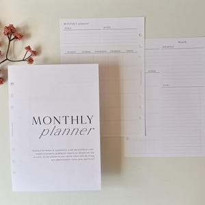 May include: A collection of white planner inserts featuring the text "MONTHLY planner" in black. The pages include sections for goals, notes, and a monthly calendar. A sprig of dried flowers is in the upper left corner.