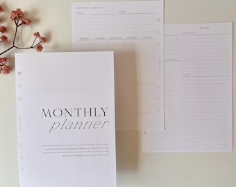 May include: A set of white planner pages with the words "MONTHLY planner" printed in black. The pages include sections for goals, notes, and a monthly calendar. A sprig of dried flowers is in the upper left corner.