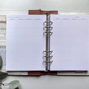 Puede incluir: Planificador mensual abierto con interior blanco, con un dise&ntilde;o de cuadr&iacute;cula para la programaci&oacute;n. El planificador est&aacute; dentro de una carpeta de cuero blanco y marr&oacute;n con anillos plateados. El lado izquierdo tiene bolsillos y una tarjeta peque&ntilde;a.