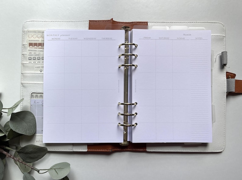 Puede incluir: Planificador mensual abierto con interior blanco, con un dise&ntilde;o de cuadr&iacute;cula para la programaci&oacute;n. El planificador est&aacute; dentro de una carpeta de cuero blanco y marr&oacute;n con anillos plateados. El lado izquierdo tiene bolsillos y una tarjeta peque&ntilde;a.
