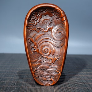 Puede incluir: Una pieza decorativa de madera tallada con un acabado marrón oscuro. El diseño presenta olas y nubes arremolinadas, creando una superficie dinámica y texturizada. La pieza tiene una forma ovalada alargada.