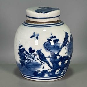May include: A white and blue ceramic jar with a lid. The jar features a hand-painted design of birds and floral elements in shades of blue. The jar is round with a lid that fits snugly on top. The design is reminiscent of traditional Asian pottery.
