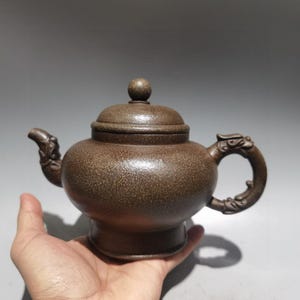 May include: A brown, speckled clay teapot with a rounded body, a lid featuring a small spherical knob, and a handle and spout both decorated with intricate, carved dragon-like motifs. The surface has a textured, matte finish throughout.