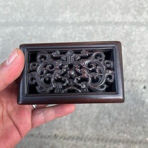 May include: A rectangular, dark brown wooden box with an intricately carved, openwork design. The carving features swirling patterns and stylized floral or foliate motifs. The box is held in a hand, showcasing the detailed craftsmanship.