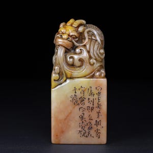 May include: A carved stone seal featuring a mythical creature with intricate details. The seal is primarily beige and yellow, with carved Chinese characters on the rectangular base. The creature has a detailed mane and a long tongue.