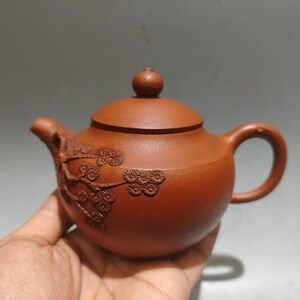 May include: A small, reddish-brown ceramic teapot with a lid and handle. The teapot features an embossed floral design on the side. The spout and handle are curved. The teapot is held in a hand.