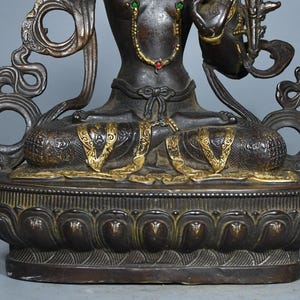 Op de afbeelding: Een donkerbronskleurig metalen standbeeld van een zittende figuur in een meditatieve houding, met goudkleurige accenten op de benen en taille, versierd met kleine rode en groene ingelegde stenen, op een gedetailleerde lotusvormige voet.