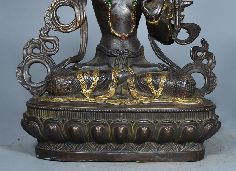 Op de afbeelding: Een donkerbronskleurig metalen standbeeld van een zittende figuur in een meditatieve houding, met goudkleurige accenten op de benen en taille, versierd met kleine rode en groene ingelegde stenen, op een gedetailleerde lotusvormige voet.