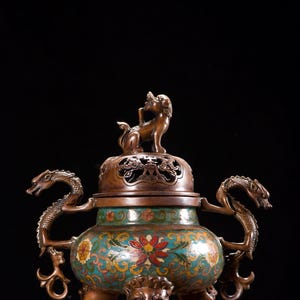 Puede incluir: Un quemador de incienso antiguo de bronce con un diseño detallado. El quemador presenta una tapa con una pequeña estatua en la parte superior, asas en forma de dragón y patrones florales y geométricos en verde azulado, rojo y amarillo. Se apoya en tres patas con forma de león.
