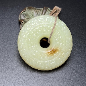 Puede incluir: Un colgante circular de piedra verde pálido con intrincados patrones tallados y un orificio central. Un cordel fino de color marrón claro atraviesa la abertura. Detrás del colgante hay un elemento decorativo metálico oscuro con forma de hoja.