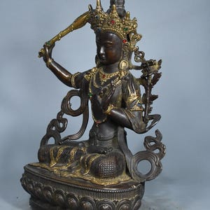 Op de afbeelding: Een donkerbronskleurig standbeeld van een zittende godheid, versierd met een gouden kroon en sieraden met kleine ingelegde rode, blauwe en groene stenen. De figuur houdt een zwaard in de rechterhand en zit op een voetstuk in lotusvorm.