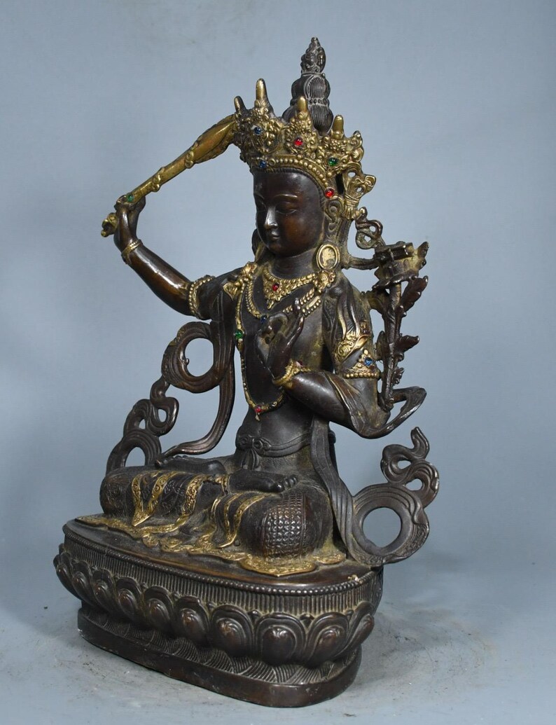 Op de afbeelding: Een donkerbronskleurig standbeeld van een zittende godheid, versierd met een gouden kroon en sieraden met kleine ingelegde rode, blauwe en groene stenen. De figuur houdt een zwaard in de rechterhand en zit op een voetstuk in lotusvorm.