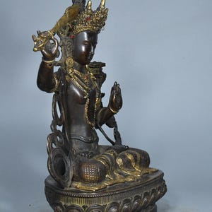 Op de afbeelding: Een donker bronskleurig standbeeld van een zittende godheid met goudkleurige accenten op de kroon, sieraden en kleding. Het figuur is versierd met kleine ingelegde rode en blauwe stenen, houdt een zwaard in de rechterhand en zit op een lotusvoet.