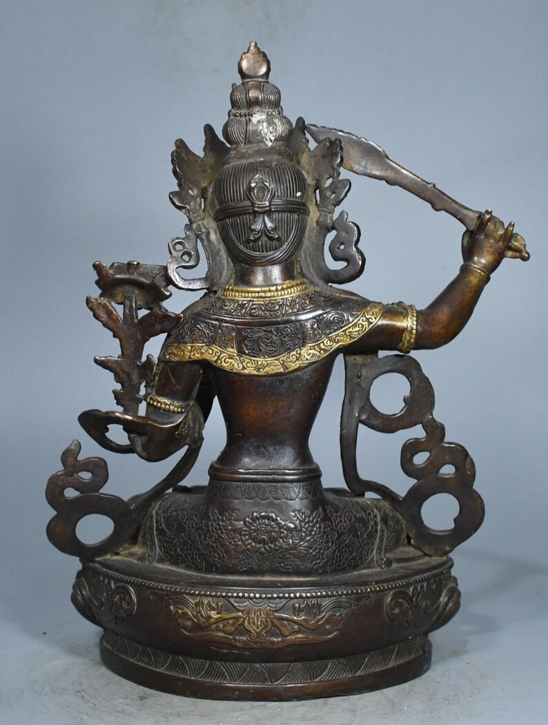 Op de afbeelding: Een bronskleurig metalen standbeeld van een godheid, gezien vanaf de achterkant. Het figuur heeft een ingewikkelde hoofdtooi, een opgeheven arm met een zwaard en een tweede arm die een bloemstengel vasthoudt. De basis en schouders hebben gouden accenten.
