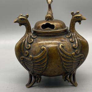 Könnte beinhalten: Ein bronzener Räuchergefäß mit Deckel und drei Beinen. Der Brenner hat zwei vogelartige Figuren auf jeder Seite und eine kleine Tierfigur auf dem Deckel. Der Brenner hat ein gealtertes Aussehen.