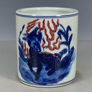 May include: A cylindrical ceramic brush pot featuring a white base with blue and orange hand-painted details. The design depicts a mythical creature with dark blue scales surrounded by orange flame-like motifs and blue foliage on a white background.