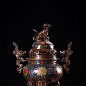 May include: An ornate, antique bronze incense burner with a detailed floral and dragon design. The blue and gold vessel features a small animal figure on top and dragon handles. The burner rests on three legs.