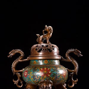 Peut inclure: Un brûleur d'encens en bronze orné avec un design détaillé. Le récipient présente un motif turquoise et floral, des poignées en forme de dragon et une petite figurine de lion sur le couvercle. Le brûleur repose sur trois pieds en forme de tête de lion.