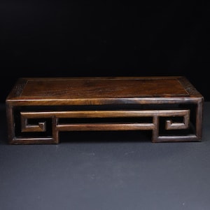 May include: A rectangular dark brown wooden display stand featuring a smooth flat top surface and a base with intricate geometric scrollwork patterns on both sides. The stand is constructed with a sturdy frame and a polished wood grain finish.