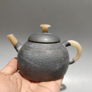 May include: A small, round, dark grey stone teapot featuring a smooth, matte finish. The teapot includes a light beige, translucent spout, a matching curved handle, and a circular lid topped with a small, light beige, cylindrical knob.