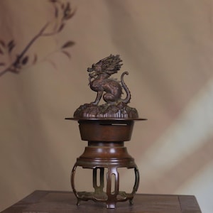 May include: A bronze-colored metal incense burner featuring a sculpted dragon figure perched atop the lid. The vessel has a cylindrical body with a wide rim and rests on a base with four curved, open-work legs and a circular support ring.