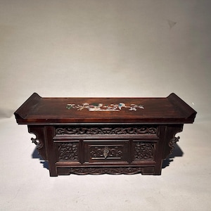 May include: A dark brown wooden altar table featuring a central drawer and intricate relief carvings on the front panels. The top surface is inlaid with a colourful mother-of-pearl design depicting birds and floral branches.