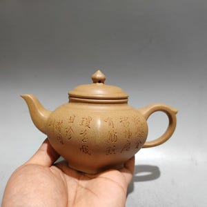 Puede incluir: Una tetera de arcilla de color marrón claro con un acabado mate texturizado, un pico curvo, un asa redondeada y una tapa con un pequeño remate puntiagudo. El cuerpo de la tetera presenta columnas verticales de caracteres chinos grabados.