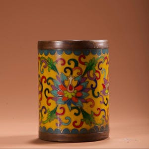 Op de afbeelding: Een cilindervormige container met een gele achtergrond en gedetailleerde bloempatronen in rood, blauw, groen en paars. De boven- en onderranden zijn donkerbrons van kleur en de container heeft een blauwe rand. Het ontwerp doet denken aan traditionele Aziatische kunst.