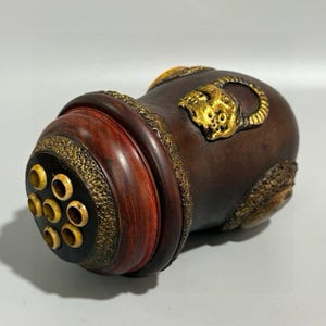 Könnte beinhalten: Dunkelbraunes, zylindrisches Dekorationsobjekt mit goldenen Akzenten. Das Objekt weist ein geschnitztes Design, ein rotes Band und eine Reihe kreisförmiger Öffnungen an einem Ende auf. Eine goldfarbene Tierfigur ziert die Seite.