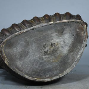 Op de afbeelding: Een donker bronskleurige metalen voet van een standbeeld met een gebogen, geribbelde rand en een plat, geborsteld oppervlak. De voet is gegraveerd met vier Chinese karakters 乾隆年製, wat duidt op een merk uit de Qianlong-periode.