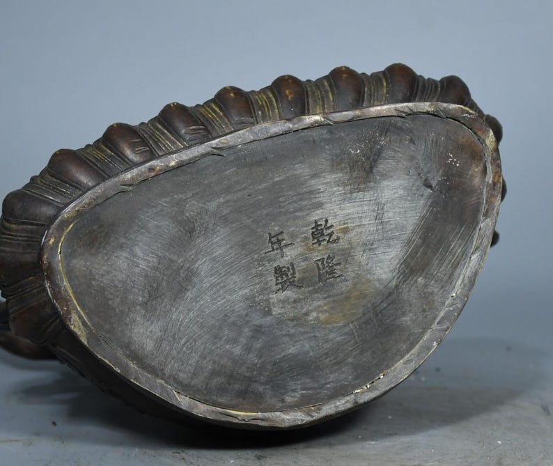 Op de afbeelding: Een donker bronskleurige metalen voet van een standbeeld met een gebogen, geribbelde rand en een plat, geborsteld oppervlak. De voet is gegraveerd met vier Chinese karakters 乾隆年製, wat duidt op een merk uit de Qianlong-periode.
