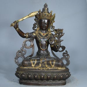 Op de afbeelding: Een donkerbronskleurig standbeeld van een zittende godheid met een gouden kroon en sieraden met kleine rode, blauwe en groene ingelegde stenen. De figuur houdt een opgeheven zwaard in de rechterhand en een lotusbloem in de linkerhand.