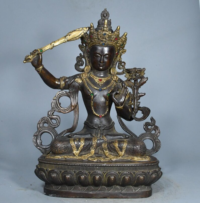 Op de afbeelding: Een donkerbronskleurig standbeeld van een zittende godheid met een gouden kroon en sieraden met kleine rode, blauwe en groene ingelegde stenen. De figuur houdt een opgeheven zwaard in de rechterhand en een lotusbloem in de linkerhand.